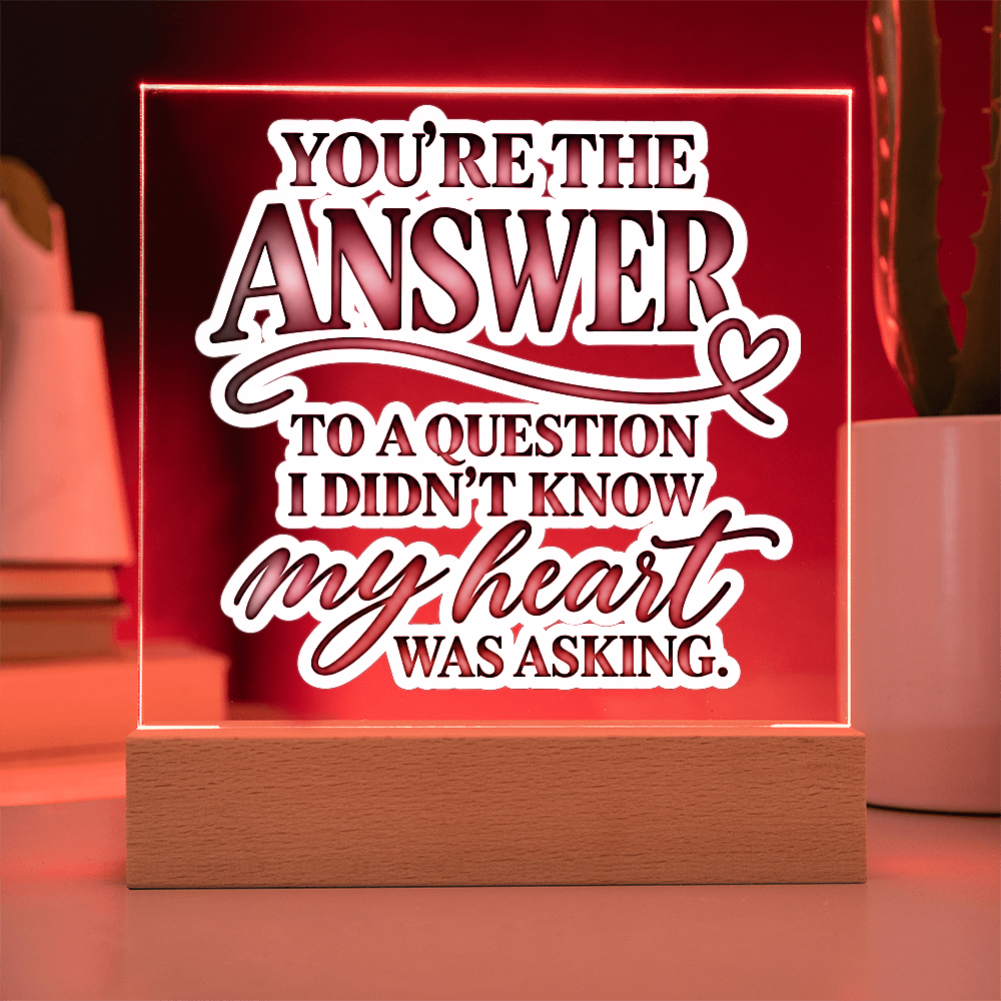 A square acrylic LED sign on a wooden base with the phrase 'YOU'RE THE ANSWER TO A QUESTION I DIDN'T KNOW MY HEART WAS ASKING.' in white and dark red lettering, glowing with a warm red light in a home setting.