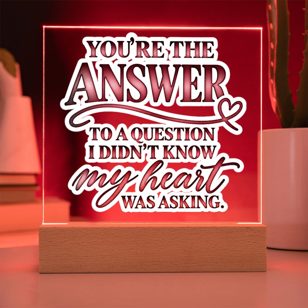 A square acrylic LED sign on a wooden base with the phrase 'YOU'RE THE ANSWER TO A QUESTION I DIDN'T KNOW MY HEART WAS ASKING.' in white and dark red lettering, glowing with a warm red light in a home setting.
