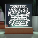 A square acrylic LED night light with a wooden base, displaying the quote 'YOU'RE THE ANSWER TO A QUESTION I DIDN'T KNOW my heart WAS ASKING.' in white and gray lettering against a dark glowing background.