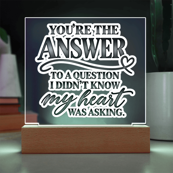 A square acrylic LED night light with a wooden base, displaying the quote 'YOU'RE THE ANSWER TO A QUESTION I DIDN'T KNOW my heart WAS ASKING.' in white and gray lettering against a dark glowing background.