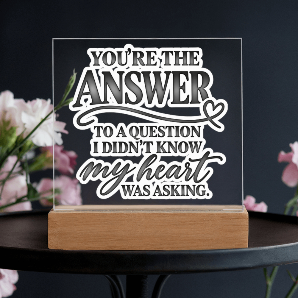 A clear acrylic LED sign with white laser-cut text reading 'YOU'RE THE ANSWER TO A QUESTION I DIDN'T KNOW my heart WAS ASKING.' displayed on a light wood base, set on a dark table with blurred pink flowers in the background.