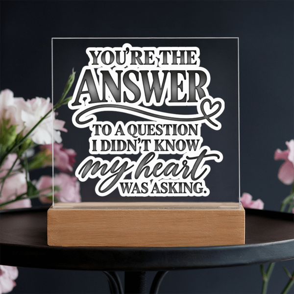 A clear acrylic LED sign with white laser-cut text reading 'YOU'RE THE ANSWER TO A QUESTION I DIDN'T KNOW my heart WAS ASKING.' displayed on a light wood base, set on a dark table with blurred pink flowers in the background.