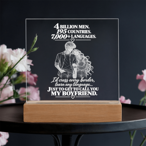 A rectangular acrylic plaque on a light wood base, engraved with a couple embracing and romantic text about counting languages and countries. The plaque is displayed on a dark table with blurred pink flowers in the background.