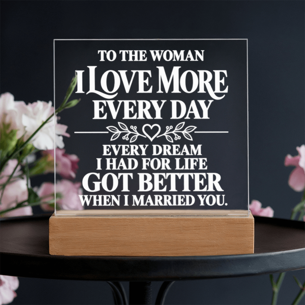 A rectangular acrylic plaque with white engraved text reading 'TO THE WOMAN I LOVE MORE EVERY DAY EVERY DREAM I HAD FOR LIFE GOT BETTER WHEN I MARRIED YOU.' displayed on a light wood base on a dark table, with blurred pink flowers in the background.