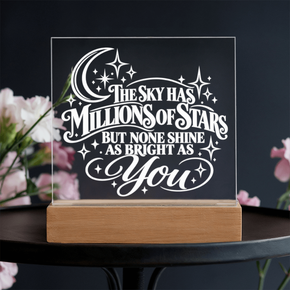 A clear acrylic LED light sign on a wooden base, featuring the quote 'THE SKY HAS MILLIONS OF STARS BUT NONE SHINE AS BRIGHT AS You' in white lettering. The sign is displayed on a dark table with blurred pink flowers in the background.