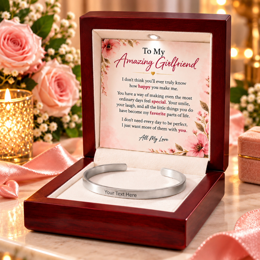A personalized silver cuff bracelet displayed inside an open dark wood gift box with a floral message card. The box is set on a reflective surface with pink roses, a lit candle, and soft bokeh lights in the background.