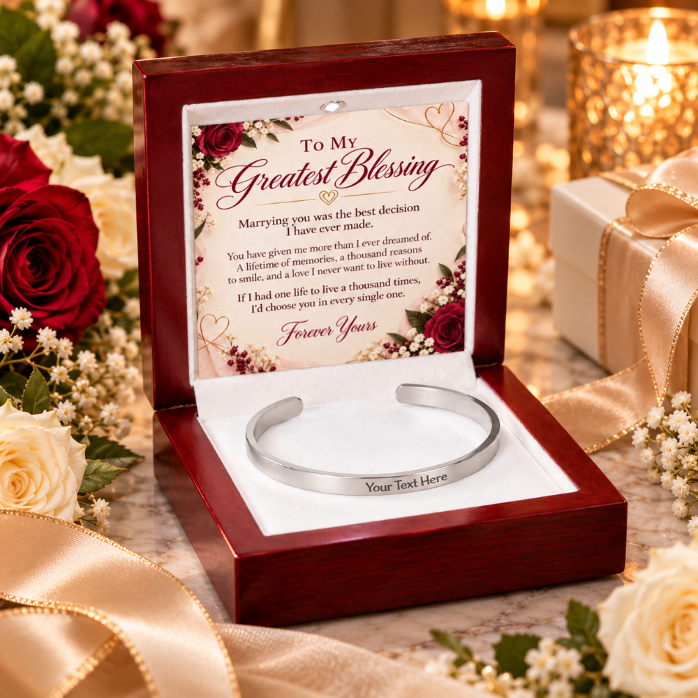 A silver engraved cuff bracelet presented in an open dark wood gift box with a floral message card inside, surrounded by roses, white flowers, and a lit candle