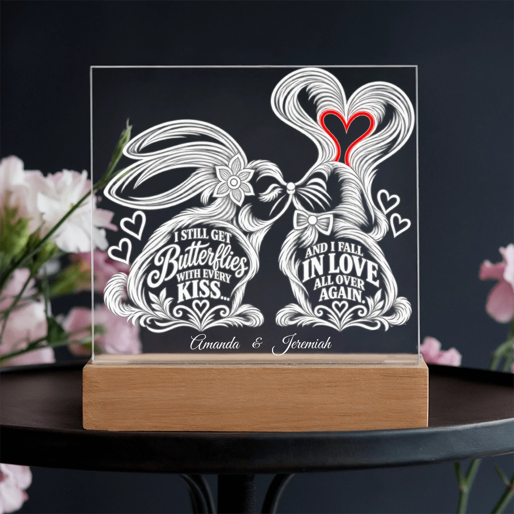 Personalized acrylic light box on a wooden base, featuring white line-art butterflies with hearts and quotes, with a red heart accent, displayed on a dark table with blurred pink flowers.