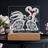 Personalized acrylic light box on a wooden base, featuring white line-art butterflies with hearts and quotes, with a red heart accent, displayed on a dark table with blurred pink flowers.