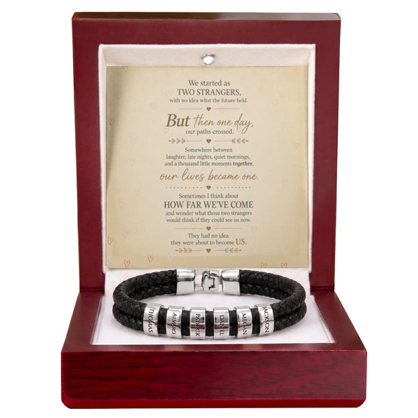 A black braided cord bracelet with silver letter beads displayed inside an open reddish-brown gift box. A cream-colored sentimental poem card is mounted behind the bracelet.