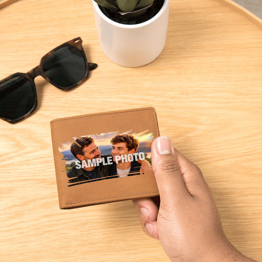 A hand holds a brown leather wallet with a custom sample photo printed on the front, set on a light wood table with sunglasses and a potted plant in the background.