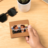 A hand holds a brown leather wallet with a custom sample photo printed on the front, set on a light wood table with sunglasses and a potted plant in the background.