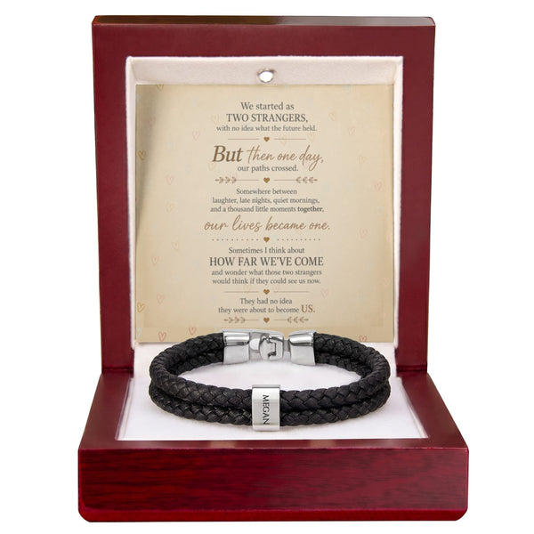 A black braided leather bracelet with a silver-toned clasp, presented inside an open reddish-brown wooden gift box. The box contains a cream-colored card with a romantic poem titled 'How Far We've Come'.
