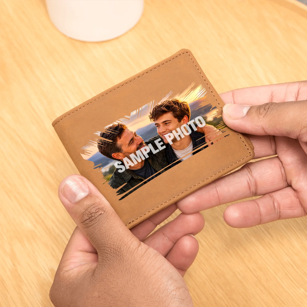 A tan leather bifold wallet being held by two hands over a light wood tabletop. The front of the wallet has a printed sample photo of two men with the text 'SAMPLE PHOTO' overlaid, indicating a customizable photo print feature.