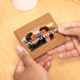 A tan leather bifold wallet being held by two hands over a light wood tabletop. The front of the wallet has a printed sample photo of two men with the text 'SAMPLE PHOTO' overlaid, indicating a customizable photo print feature.
