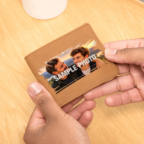 A tan leather bifold wallet being held by two hands over a light wood tabletop. The front of the wallet has a printed sample photo of two men with the text 'SAMPLE PHOTO' overlaid, indicating a customizable photo print feature.