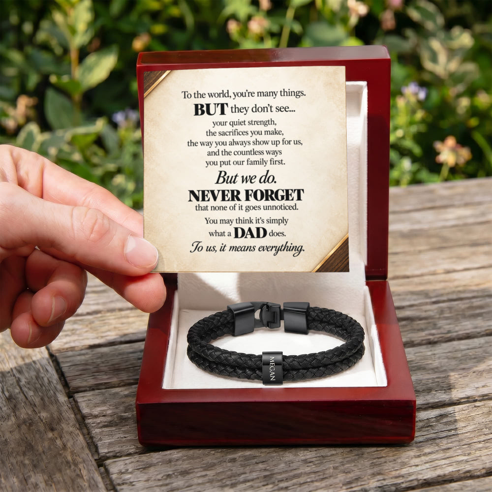 A hand holds a personalized black braided cord bracelet inside an open red presentation box, with a printed message card displayed above it on a wooden table outdoors.