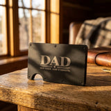 A black metal slim wallet with a cutout for a card, featuring the text 'DAD OUR ROCK. OUR STRENGTH.