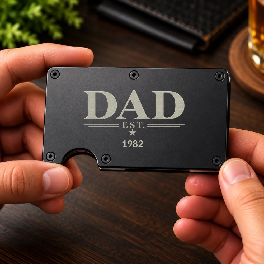 A person's hands holding a slim black metal card holder engraved with 'DAD EST. 1982' in silver lettering, set against a dark wooden table background.