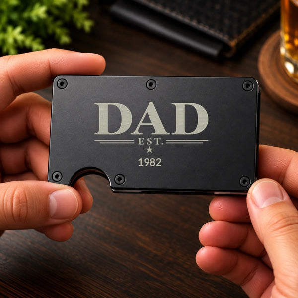 A person's hands holding a slim black metal card holder engraved with 'DAD EST. 1982' in silver lettering, set against a dark wooden table background.