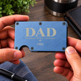 A person holds a slim blue metal card holder with 'DAD EST. 1982' printed on it, shown over a wooden desk background with a pen and notebook nearby.