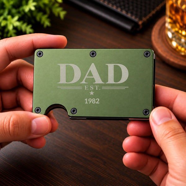 A person's hands holding a slim, olive green metal card wallet engraved with 'DAD EST. 1982' on a dark wooden table background.