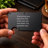 Black personalized metal wallet for son held in two hands, showing the engraved message on the front.