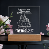 A clear acrylic plaque on a light wood base, engraved with a romantic message and couple illustration, displayed on a dark table with blurred pink flowers in the background.