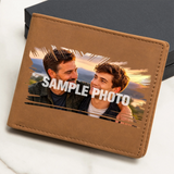 A brown faux-leather bifold wallet shown at an angle on a light marble surface with a dark box behind it. The front has a printed sample photo of two men with the text 'SAMPLE PHOTO' overlaid in white.