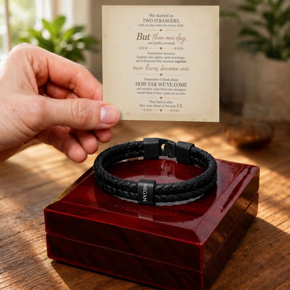 A black braided men's bracelet with a black metal clasp and a small silver-toned tag reading 'LOVE' is displayed on a glossy red presentation box. A hand holds a printed sentimental message card above the box, with a warm, softly blurred indoor background.