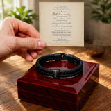 A black braided men's bracelet with a black metal clasp and a small silver-toned tag reading 'LOVE' is displayed on a glossy red presentation box. A hand holds a printed sentimental message card above the box, with a warm, softly blurred indoor background.