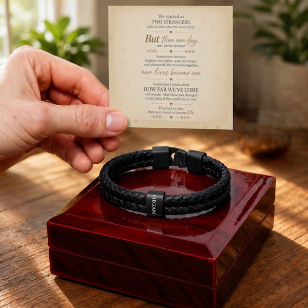 A black braided men's bracelet with a black metal clasp and a small silver-toned tag reading 'LOVE' is displayed on a glossy red presentation box. A hand holds a printed sentimental message card above the box, with a warm, softly blurred indoor background.