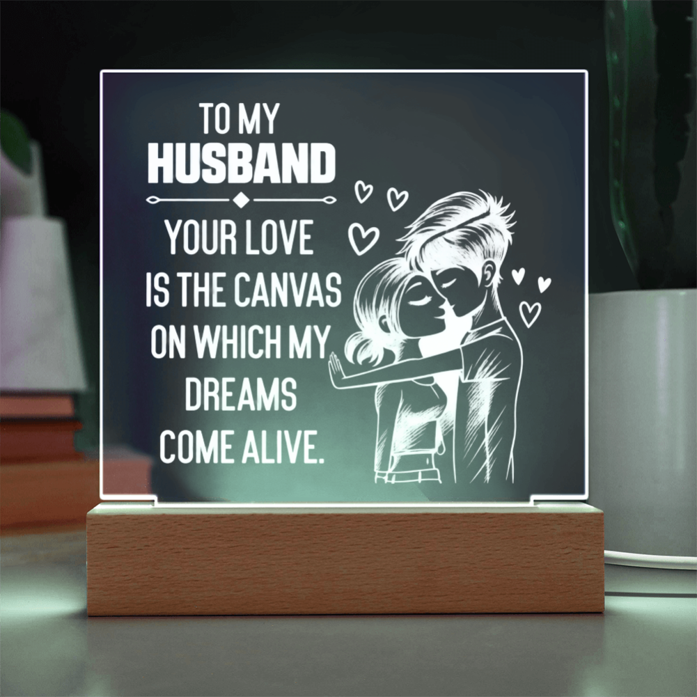 A rectangular acrylic LED light sign on a wooden base, displaying a romantic message for a husband: 'TO MY HUSBAND YOUR LOVE IS THE CANVAS ON WHICH MY DREAMS COME ALIVE,' with a white illustration of a couple embracing and small hearts, shown on a tabletop.