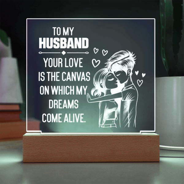 A rectangular acrylic LED light sign on a wooden base, displaying a romantic message for a husband: 'TO MY HUSBAND YOUR LOVE IS THE CANVAS ON WHICH MY DREAMS COME ALIVE,' with a white illustration of a couple embracing and small hearts, shown on a tabletop.