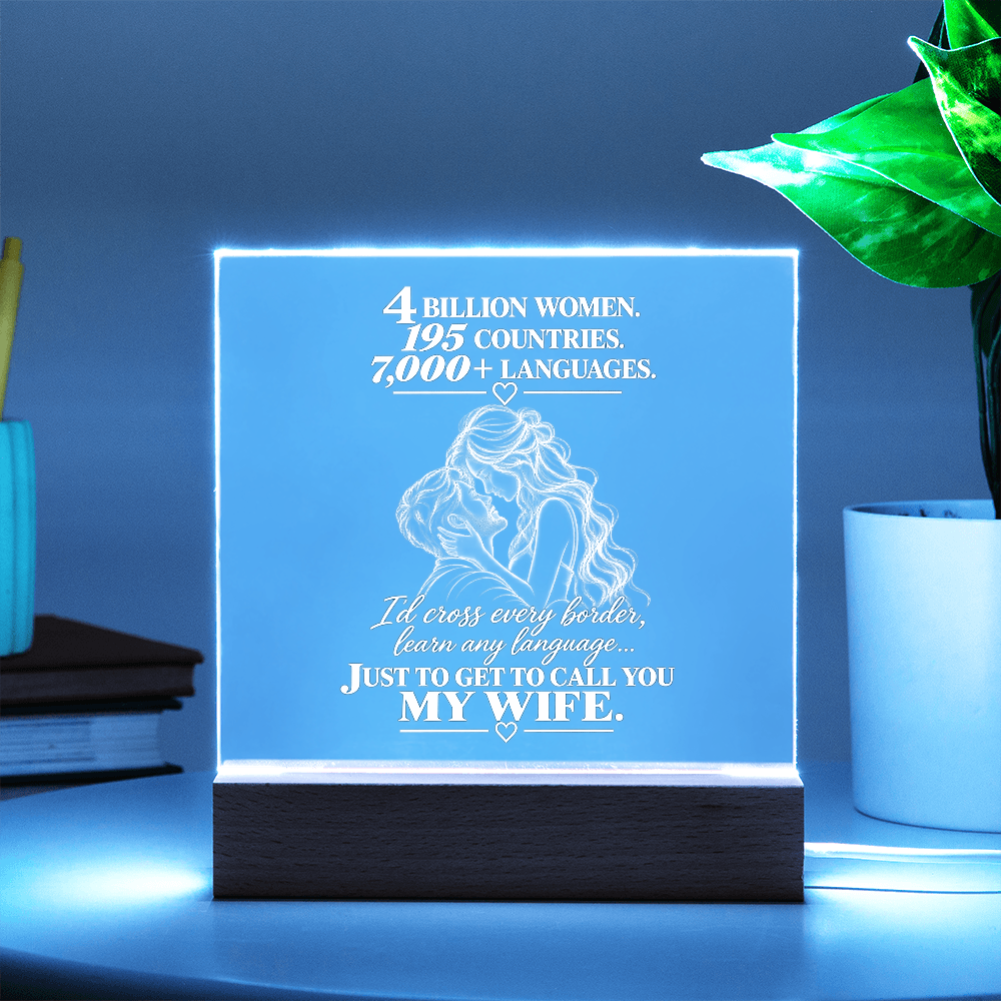 A glowing acrylic LED cube lamp on a dark wooden base, displaying a romantic quote and an illustration of a couple. The lamp is styled on a table next to a potted plant and books, with a cool blue ambient glow.