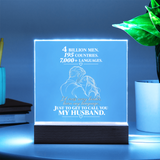A glowing acrylic LED cube lamp on a dark wooden base, displaying a romantic quote and an illustration of a couple. The lamp is styled on a table next to a potted plant and books, with a cool blue ambient glow.