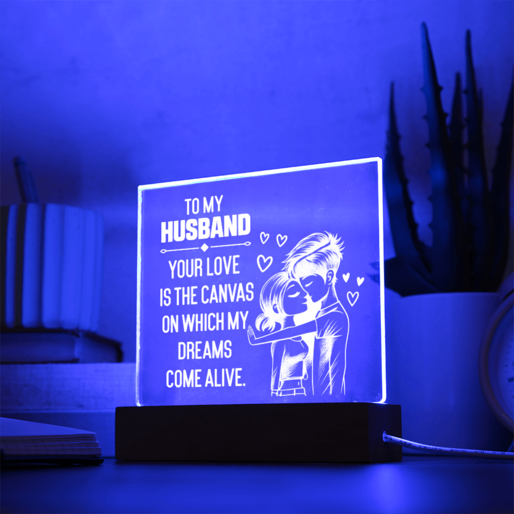 A rectangular acrylic LED night light on a black base, glowing with a blue light and featuring engraved white text that reads, 'TO MY HUSBAND YOUR LOVE IS THE CANVAS ON WHICH MY DREAMS COME ALIVE,' alongside an illustration of a couple embracing. The lamp is placed on a tabletop in a styled room setting with books and a potted cactus in the background.