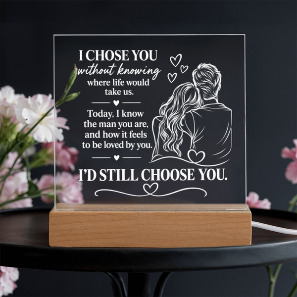 Personalized acrylic LED night light with a wooden base, featuring a romantic engraved message and illustration of a couple, displayed on a dark table with blurred pink flowers in the background.
