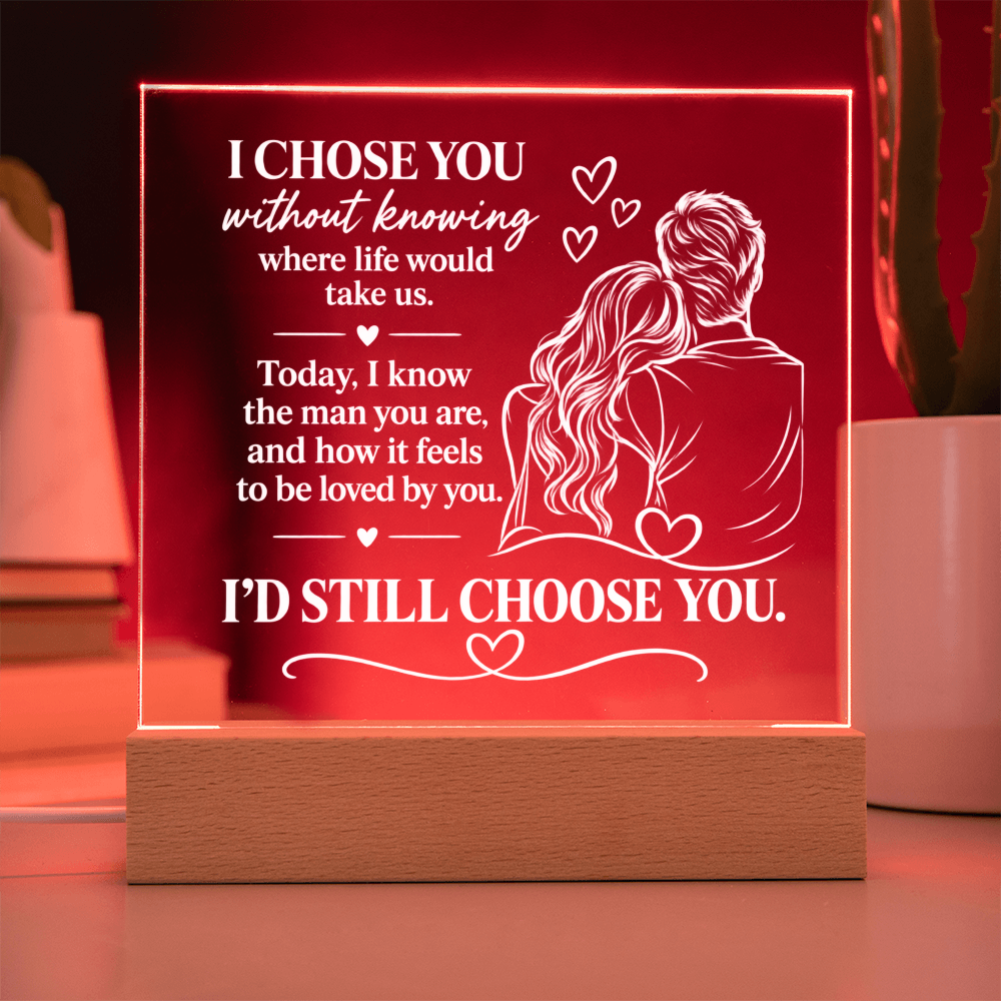 A rectangular acrylic LED light sign on a wooden base, glowing red with white engraved text and an illustration of a couple, shown on a tabletop with blurred plants in the background.