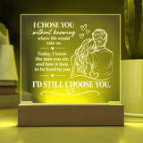 A rectangular acrylic LED night light with a wooden base, glowing with a warm yellow light. The front features a white laser-engraved illustration of a couple from behind with hearts and romantic text, set against a softly blurred home background.