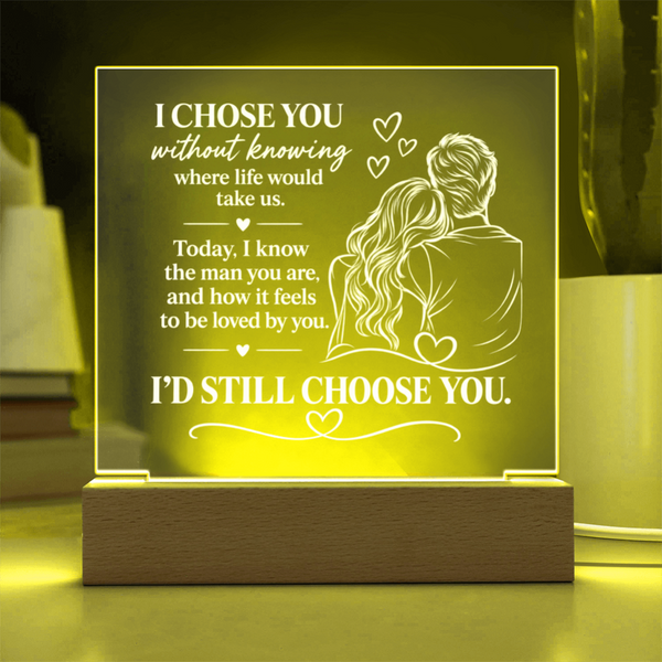 A rectangular acrylic LED night light with a wooden base, glowing with a warm yellow light. The front features a white laser-engraved illustration of a couple from behind with hearts and romantic text, set against a softly blurred home background.