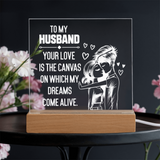 A rectangular acrylic LED light box on a wooden base, featuring a romantic quote for a husband and an illustration of a couple with hearts, displayed on a dark table with blurred pink flowers.