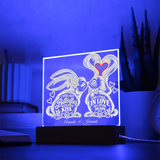 A personalized acrylic LED light box on a black base, glowing with a blue light and featuring a bunny illustration and the names 'Annelise & Jeremiah' with romantic quotes. The scene is set on a table with a blurred background of books and a plant.