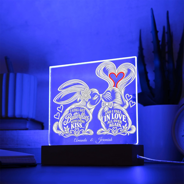 A personalized acrylic LED light box on a black base, glowing with a blue light and featuring a bunny illustration and the names 'Annelise & Jeremiah' with romantic quotes. The scene is set on a table with a blurred background of books and a plant.