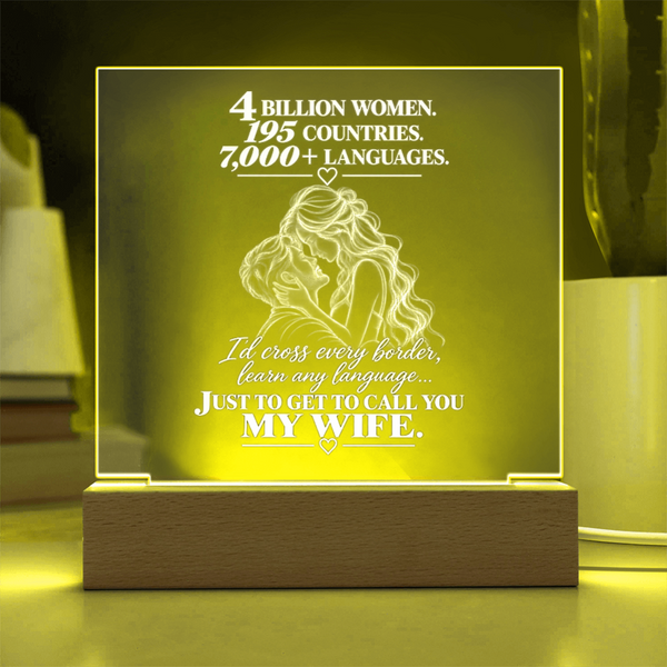 A rectangular acrylic LED night light with a wooden base, glowing with a warm yellow light. The front features a white laser-engraved illustration of a couple from behind with hearts and romantic text, set against a softly blurred home background.