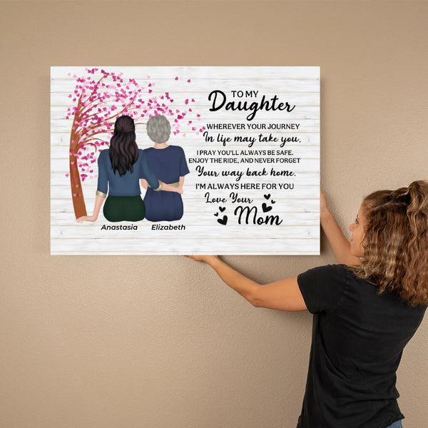 A woman holds a personalized canvas print against a beige wall. The print features a white wood-plank background with a pink cherry blossom tree, the silhouettes of two women, and a sentimental message addressed to a daughter titled 'TO MY Daughter'.