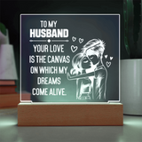 A rectangular acrylic LED light sign on a wooden base, displaying a romantic message for a husband: 'TO MY HUSBAND YOUR LOVE IS THE CANVAS ON WHICH MY DREAMS COME ALIVE' with a white illustration of a couple and small hearts, shown in a home-like setting.