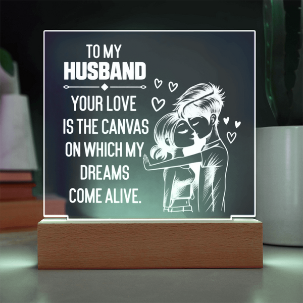 A rectangular acrylic LED light sign on a wooden base, displaying a romantic message for a husband: 'TO MY HUSBAND YOUR LOVE IS THE CANVAS ON WHICH MY DREAMS COME ALIVE' with a white illustration of a couple and small hearts, shown in a home-like setting.