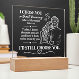 A personalized acrylic LED light plaque on a wooden block base, featuring white engraved text and an illustration of a couple, displayed on a tabletop with blurred plants and books in the background.