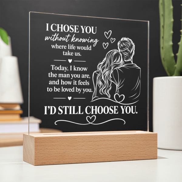 A personalized acrylic LED light plaque on a wooden block base, featuring white engraved text and an illustration of a couple, displayed on a tabletop with blurred plants and books in the background.